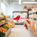 Main tenant une carte de fidélité Auchan Waaoh devant un rayon de supermarché moderne