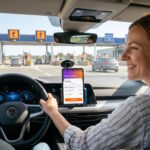 Conducteur souriant au volant regardant son application Ulys sur smartphone, avec une barrière de télépéage en arrière-plan.