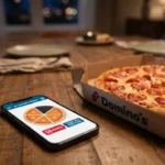 Smartphone affichant la jauge de fidélité Domino's Pizza avec 5 parts sur 6 remplies, posé à côté d'une pizza pepperoni.